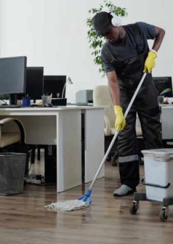 Contemporary young black man in workwear cleaning floor in openspace office in front of yellow plastic signboard with caution